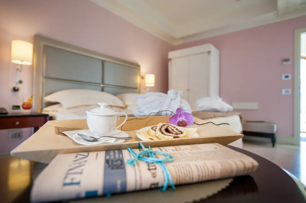 The image shows a detail of a hotel room. On a dark wooden round table is a newspaper tied with a blue ribbon. Next to it is a light wooden tray with a cup, a saucer with two slices of chocolate swirl, and a flower. In the background, you can see the bed and wardrobe.