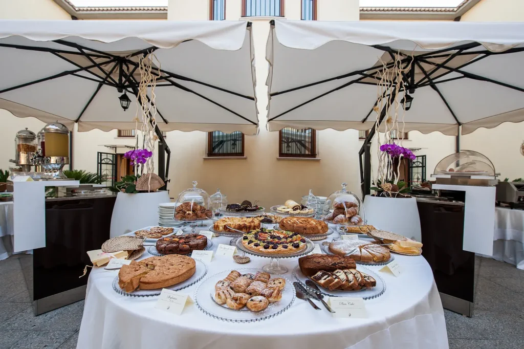 Nell'immagine la colazione dell'hotel. Al centro, sopra un tavolo con tovaglia bianca ci sono dei vassoi e alzatine con diversi prodotti per la colazione: torte a fette, biscotti, croissants. Sopra ci sono due grandi ombrelloni beige.