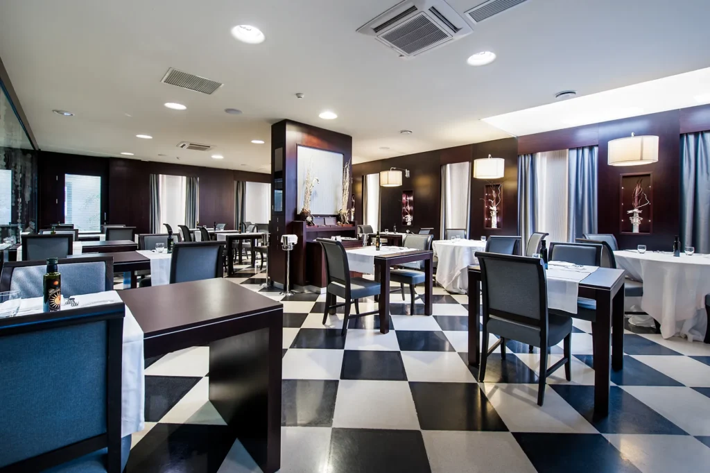 Nell'immagine la sala ristorante dell'hotel. La stanza è occupata da tavoli di legno scuro e sedie di legno scuro e velluto azzurro. Le pareti sono di legno scuro con grandi appliques e piccoli vasi decorativi in nicchie. Il pavimento è a scacchi nero e bianco.