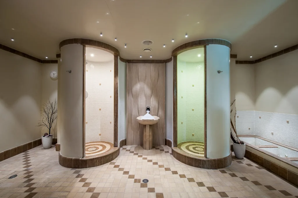 Nell'immagine la spa dell'hotel con al centro due docce in muratura tonde con luci di differenti colori. Tra le due docce c'è un lavandino con del ghiaccio. A destra si intravedono dei lavapiedi sempre in muratura. Il pavimento è composto da piccole piastrelle marrone e beige.