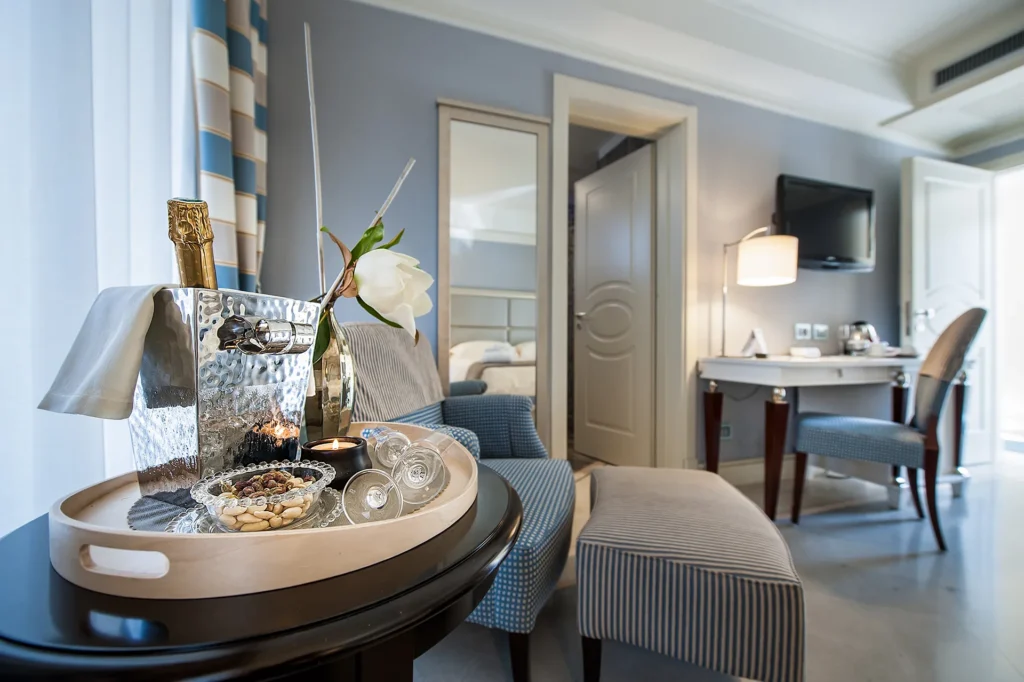 The image shows a detail of the hotel suites. On the left, atop a brown coffee table, there's a light wood tray with a small glass bowl of dried fruit, a black candle, two champagne flutes, an ice bucket containing a bottle of sparkling wine, and a fière. Behind it is an armchair with an ottoman. Behind the armchair is a full-length mirror hanging on the wall and a half-open door. To the left of the door is a desk with a chair, a table lamp, and a TV mounted on the wall.