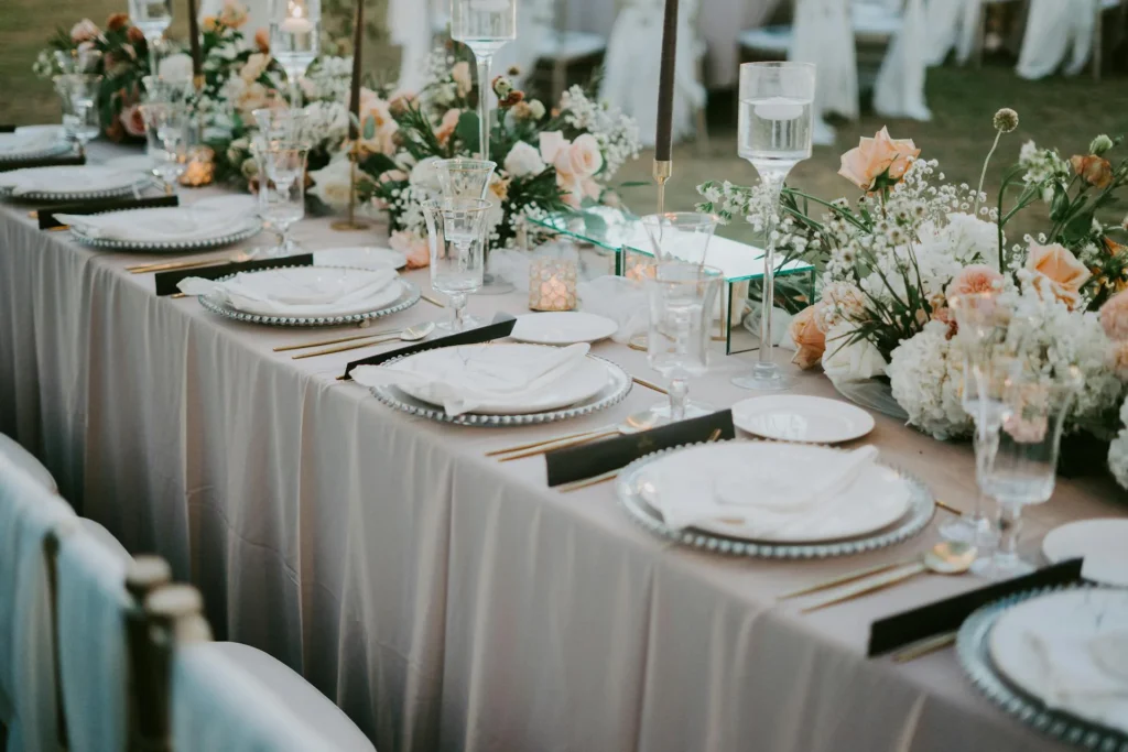 The image shows a table set for a ceremony with silver placemats, white plates, napkins, silverware, and glasses. The tablecloth is pale pink. There are candles and tall clear jars with water and floating candles, as well as floral arrangements.