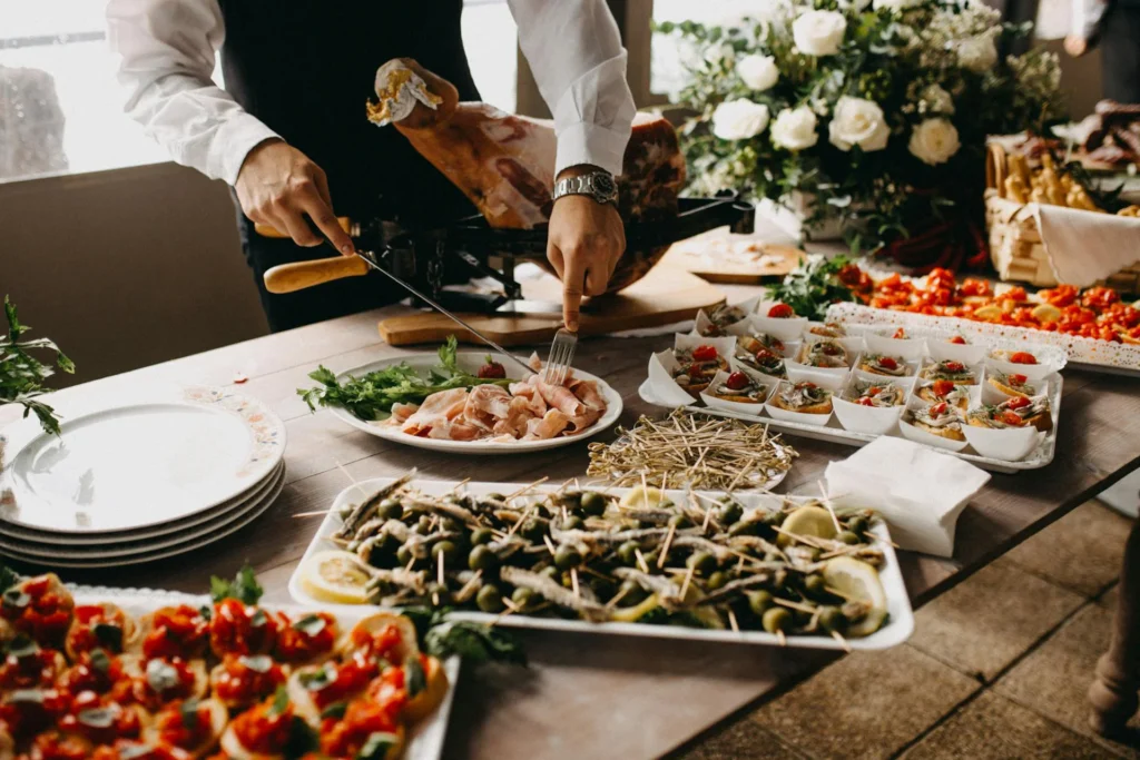 The image shows a table set with trays of various products: crostini with cherry tomatoes, anchovies, and olives, as well as various appetizers. Behind the table, a waiter is placing freshly cut slices of prosciutto on a plate. On the table to the right, there's also a vase with white roses and greenery.