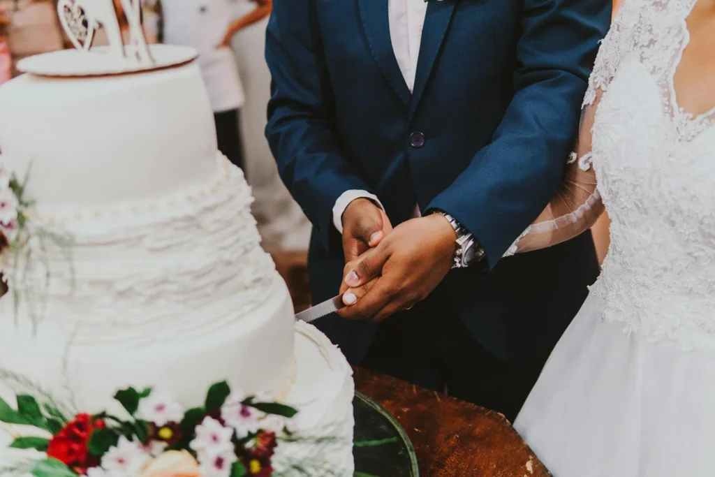 The image shows a newlywed couple holding a knife and cutting a slice of cake. The cake is tiered, white, and features flowers in the foreground.