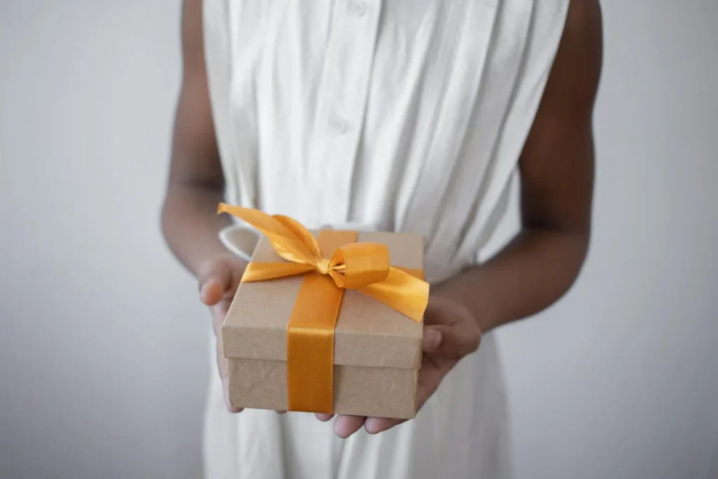 In the image, a girl in a white dress holds a gift package. The box is brown with a large orange ribbon tied in a bow.