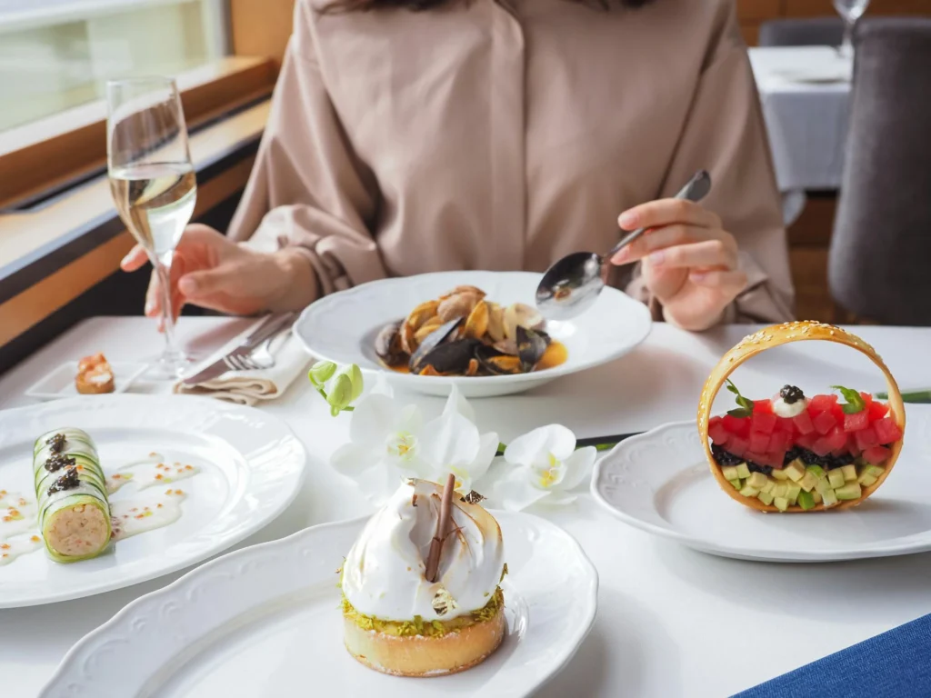 The image shows plates on a table with various dishes inside. On the left is a roll with white sauce, in the center is a meringue tartlet, on the left is a circle of pasta with diced fruit and vegetables inside, and behind it is a plate of mussels. A girl is sitting behind the table, holding a glass of prosecco in one hand and a spoon in the other, which she is bringing to the plate of mussels.