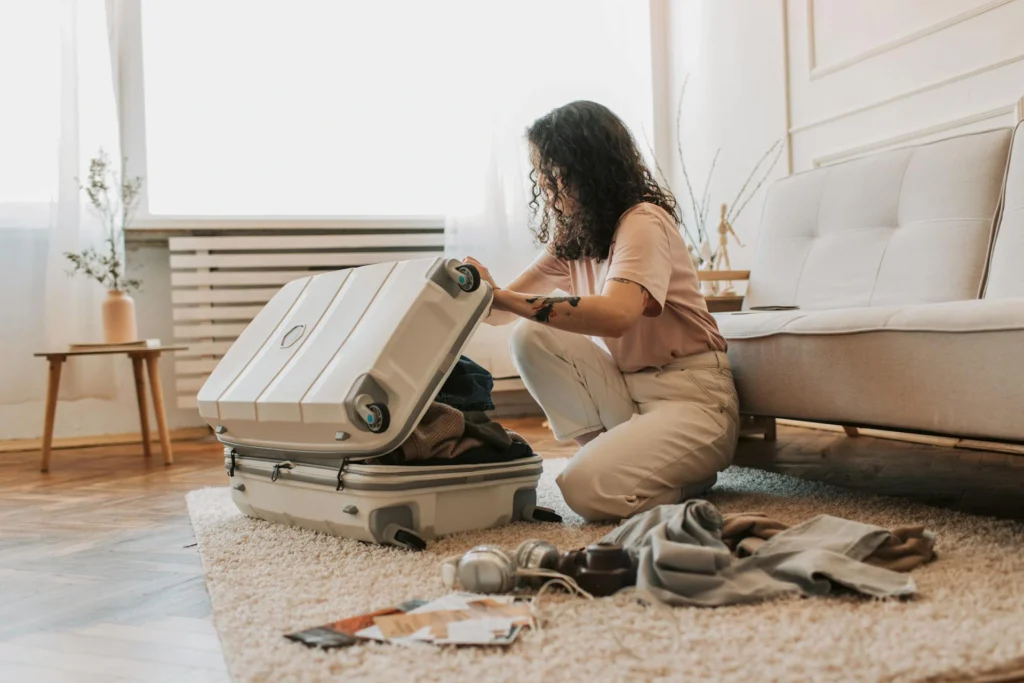 The image shows a girl kneeling on the floor on a rug, trying to close a white suitcase full of clothes. Beside her are other objects. Behind her is a sofa. In the background, you can see a low table with a vase of greenery and a window.