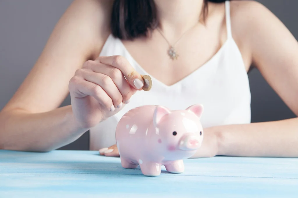 In the image, a girl inserts a coin into a pink piggy bank on a blue surface. She's wearing a white tank top and a pendant necklace around her neck.