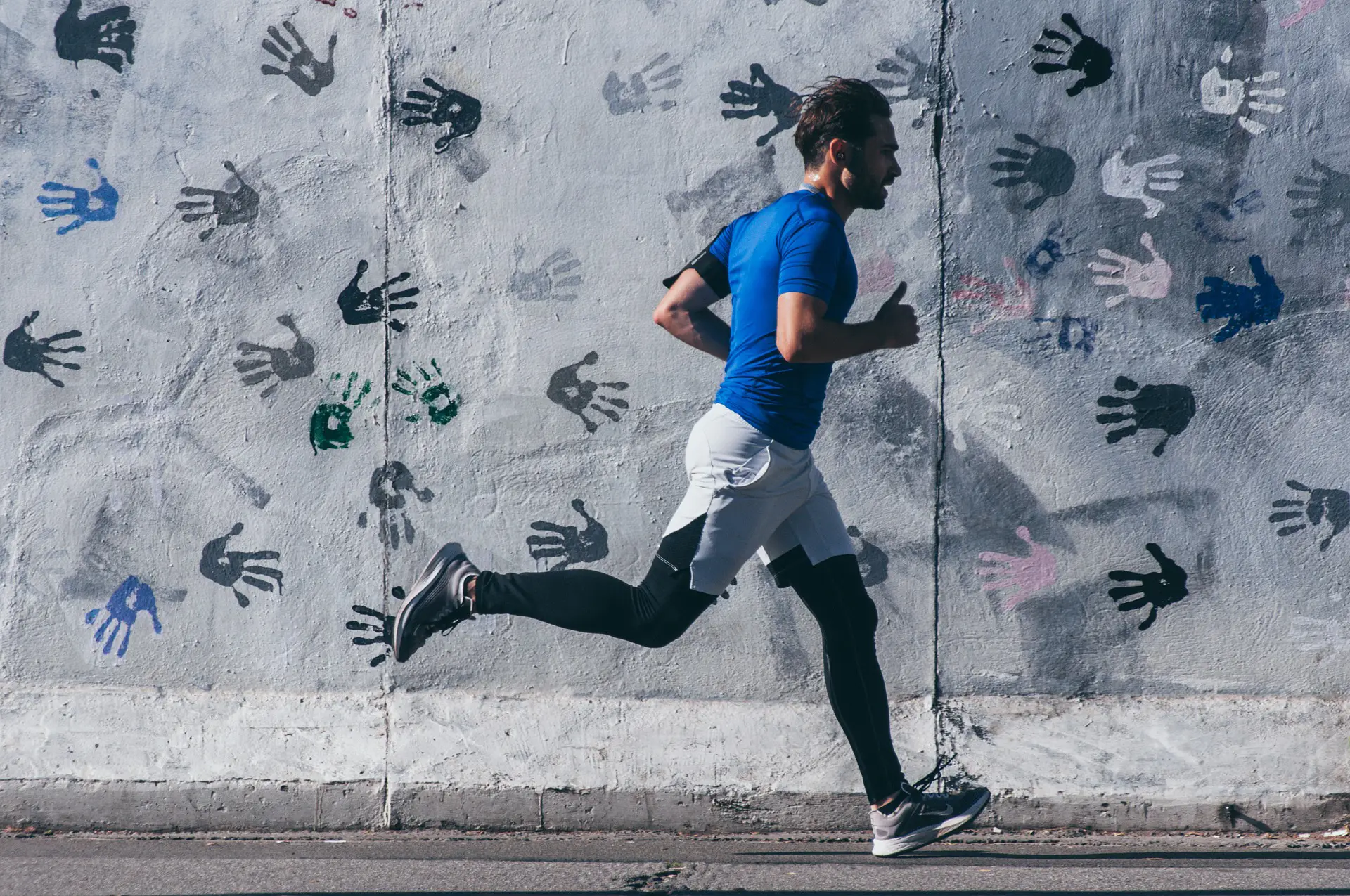 Nell'immagine c'è un ragazzo che corre. Lui indossa scarpe da running, scaldamuscoli neri, pantaloncini bianchi e una t-shirt blu. Dietro di lui c'è un muro sul quale ci sono delle impronte di mani fatte con la vernice.